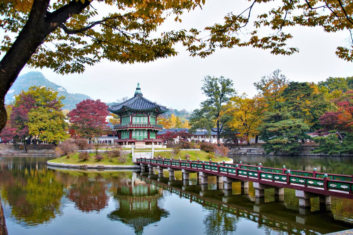 O Pavilhão Hyangwonjeong no Palácio Gyeongbokgung em Seul cercado pela folhagem de outono, com árvores em tons vibrantes de vermelho, amarelo e laranja refletidas em um lago tranquilo.