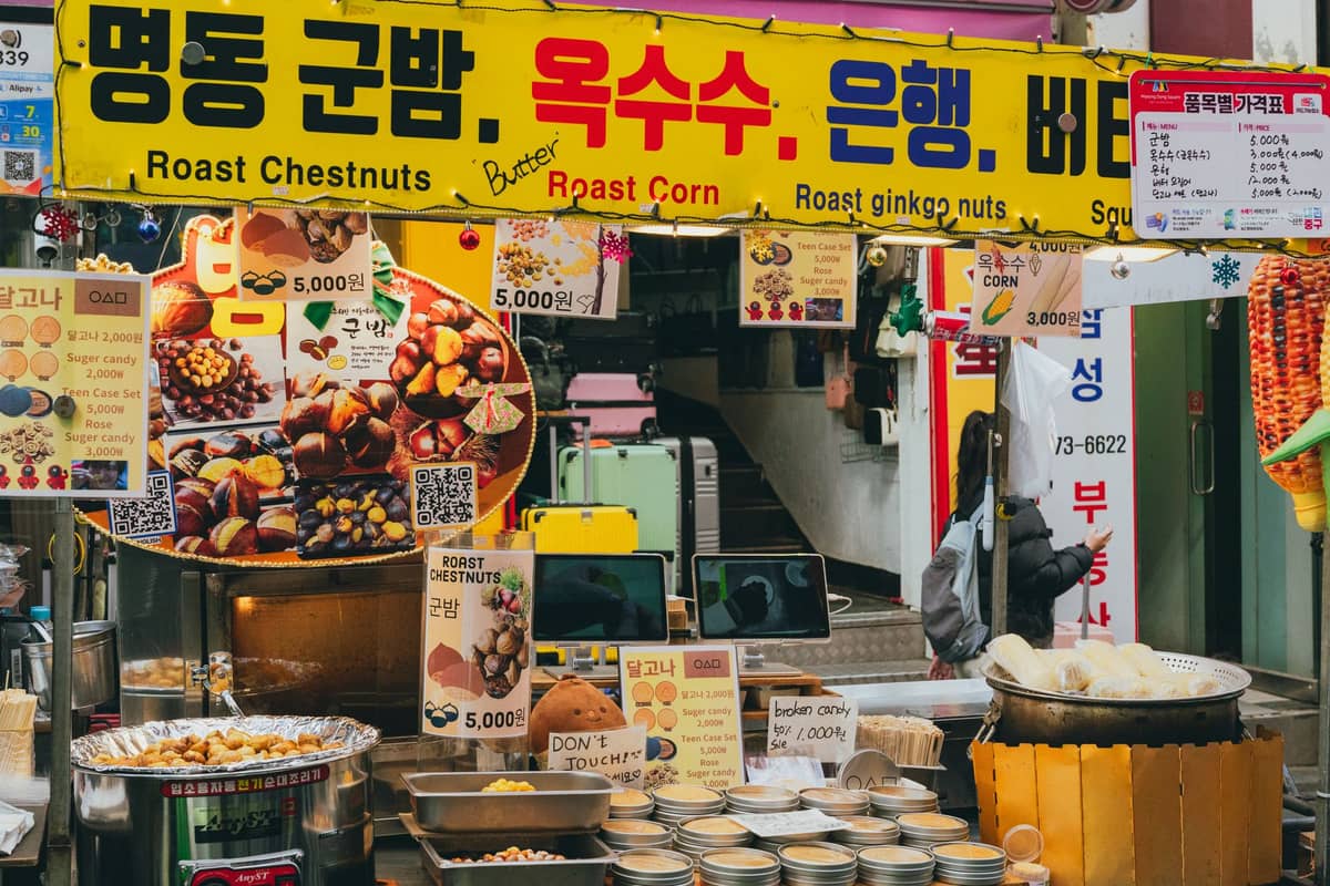 Barraca de comida de rua em Seul vendendo castanhas assadas, milho e nozes de ginkgo com placas coreanas amarelo-brilhantes e vários petiscos de mercado em exposição.