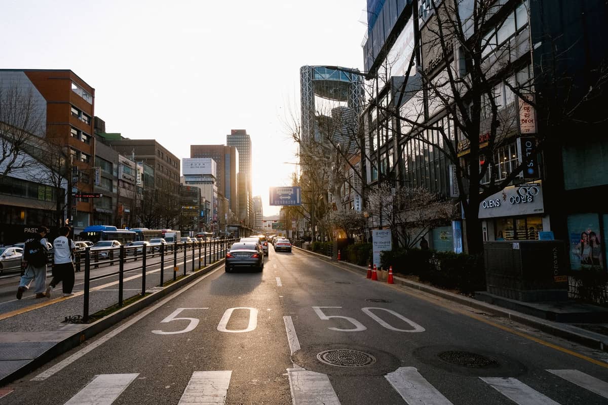 Ampla rua de Seul sob luz solar suave com edifícios modernos, árvores sem folhas, carros e alguns pedestres, capturando a atmosfera do início da primavera.