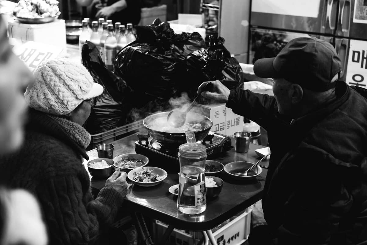 Duas pessoas com roupas de inverno comendo hot pot fumegante em uma mesa pequena em um movimentado mercado de rua coreano.