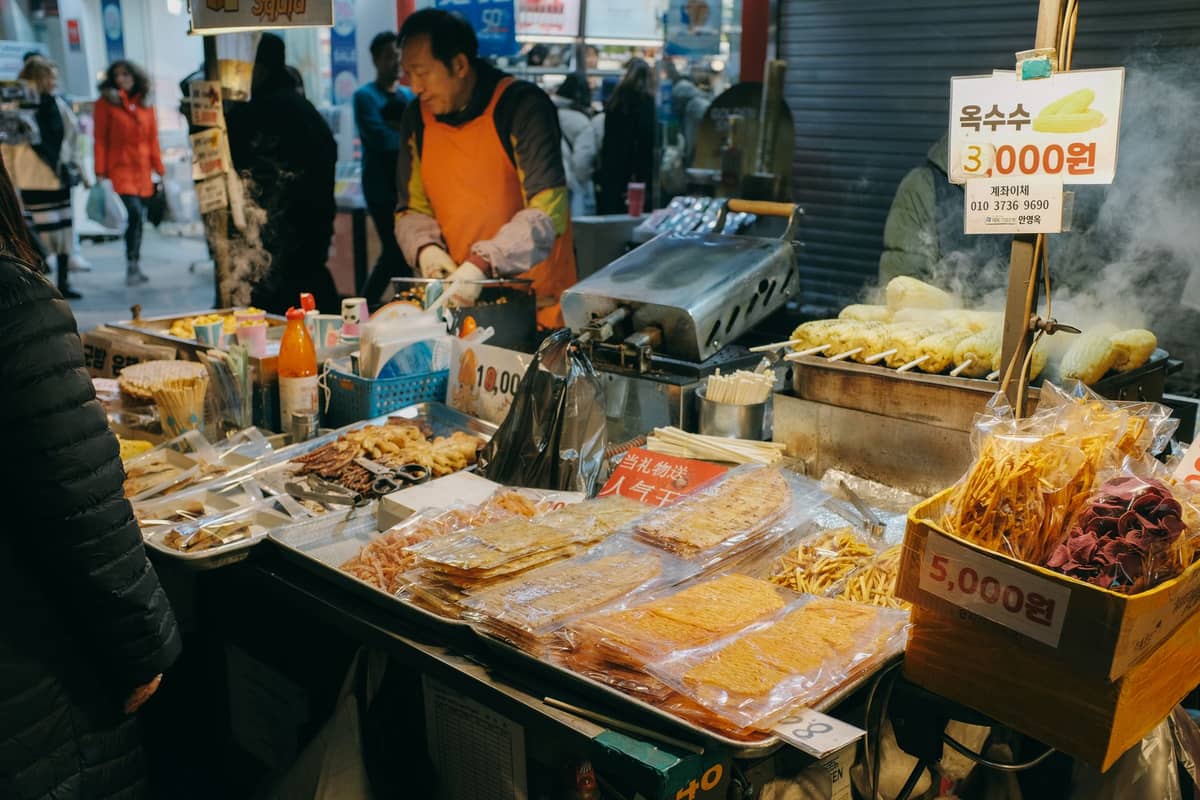 Uma movimentada barraca de mercado de rua em Seul vendendo uma variedade de petiscos, incluindo bolinhos de peixe e milho, com clientes e placas de preços escritas à mão em coreano.