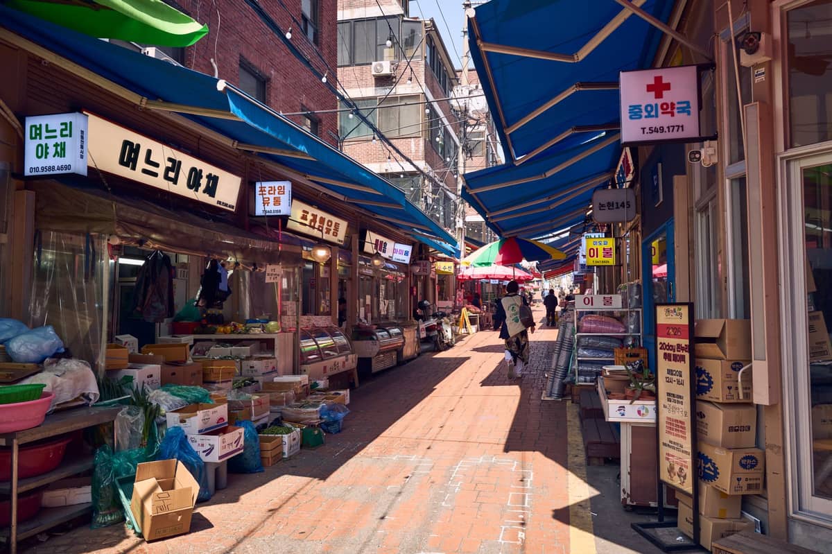 Beco externo de um mercado tradicional coreano em Seul, com toldos azuis, fachadas de lojas, produtos em exposição e letreiros em coreano.