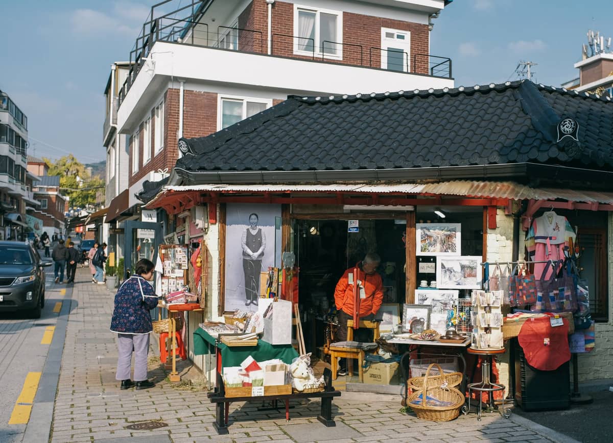 Mercado de rua em uma esquina de Seul com uma barraca vendendo artesanatos tradicionais, obras de arte, têxteis e souvenirs, com uma pessoa navegando em frente à loja.