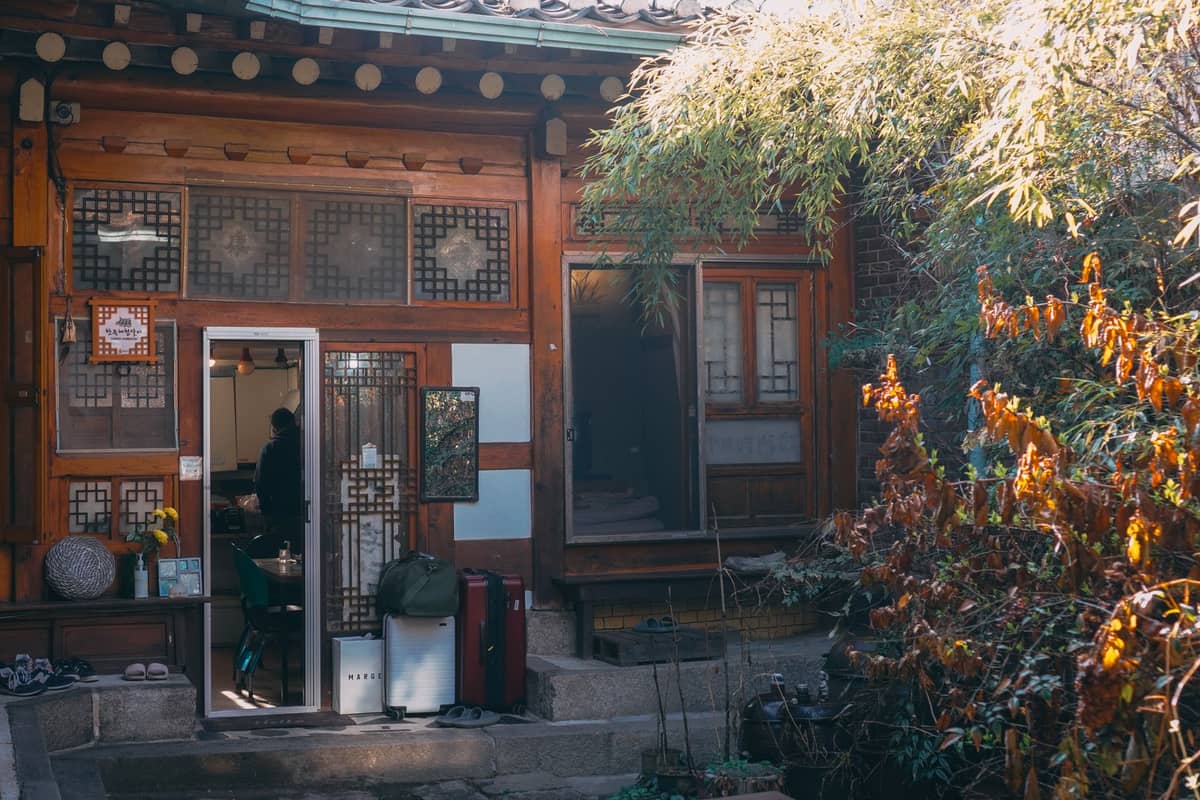 Vista do pátio de uma casa de hóspedes hanok tradicional em Seul com portas abertas, bagagens visíveis e plantas, sugerindo uma acomodação local acessível.