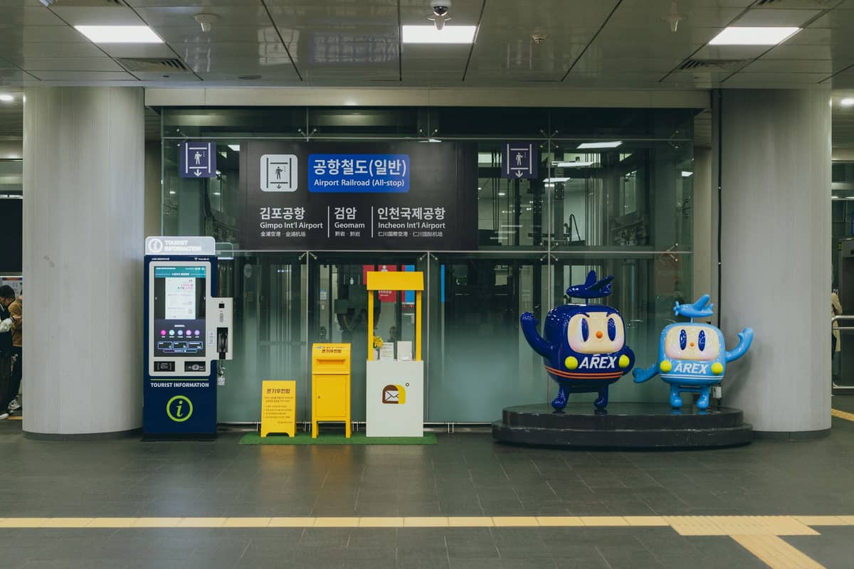Entrada para o trem aeroportuário AREX em uma estação de Seul, com máquinas de bilhetes e mascotes da AREX visíveis.