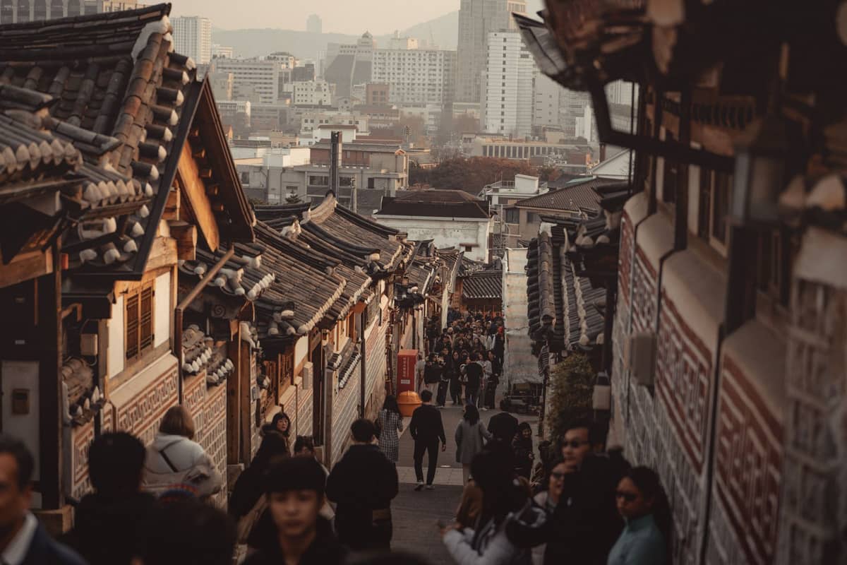 Rua da aldeia Bukchon Hanok em Seul com casas tradicionais, turistas caminhando e edifícios modernos ao fundo.