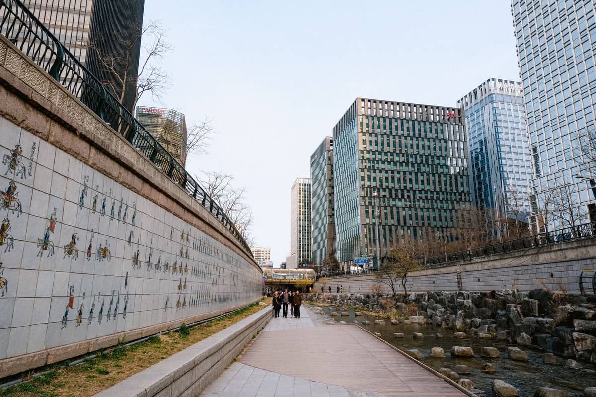 Calçadão às margens do Córrego Cheonggyecheon no centro de Seul, ladeado por modernos edifícios comerciais e azulejos decorativos nas paredes, com a vida urbana ao fundo.
