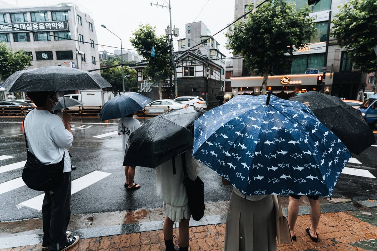Pessoas com guarda-chuvas esperando em uma esquina chuvosa em uma cidade moderna da Coreia com letreiros em hangul.