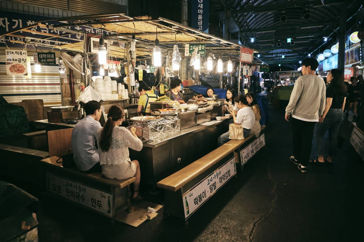 Cena noturna em um movimentado mercado de comida de rua em Seul, com pessoas sentadas em bancos comendo e vendedores preparando pratos variados sob luzes brilhantes.