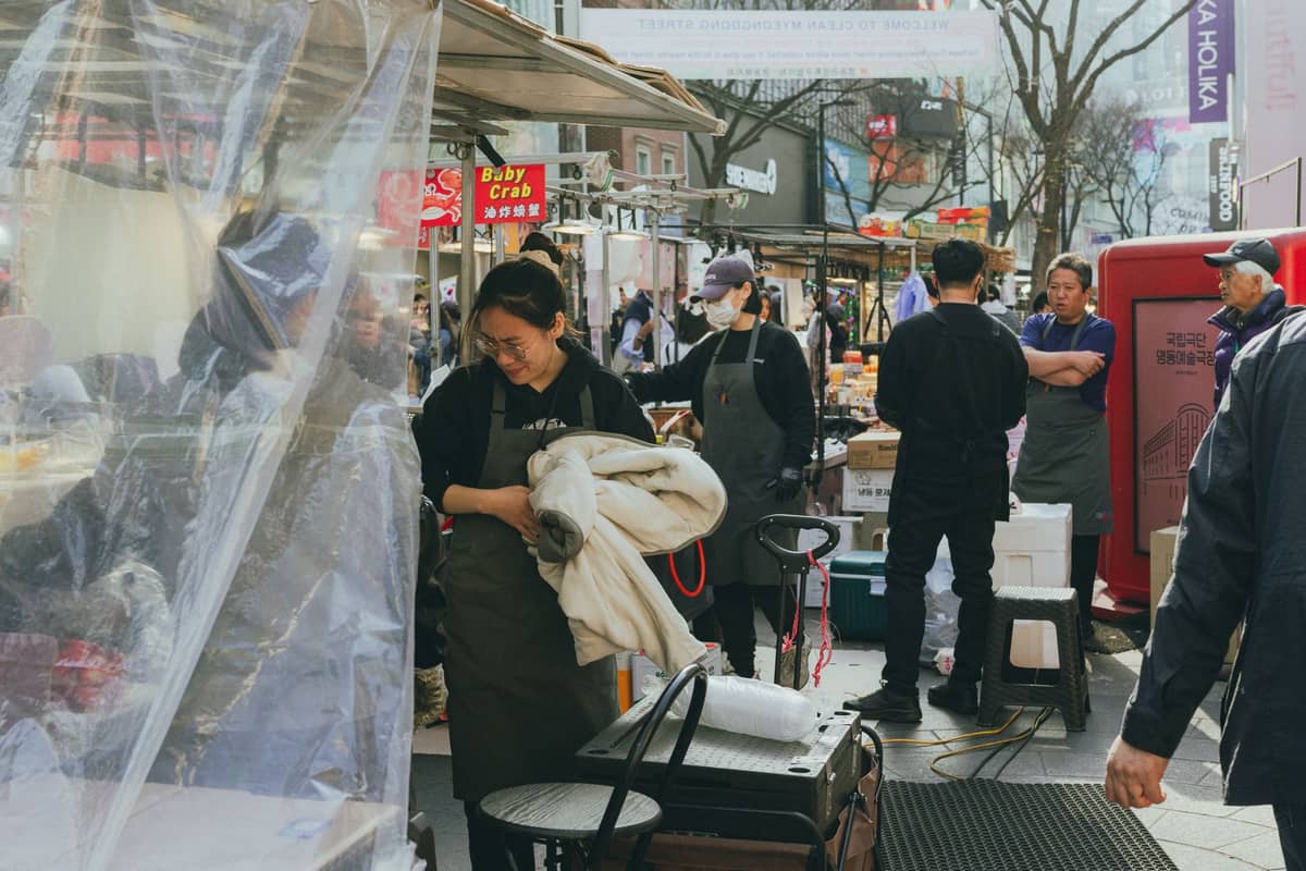 Mercado de rua ao ar livre em Seul com vendedores e barreiras climáticas de plástico, pessoas usando jaquetas e árvores do início da primavera.