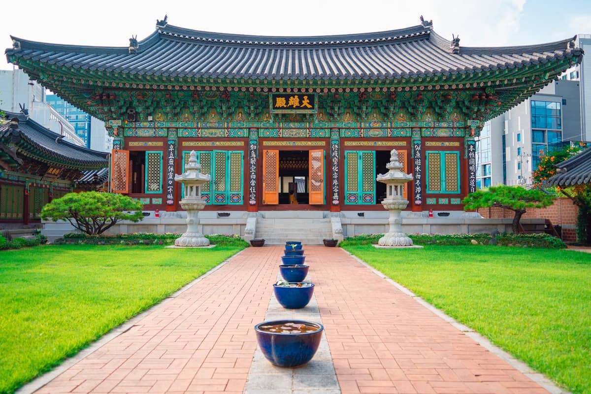Salão principal de um templo budista tradicional em Seul com ornamentada arquitetura em madeira, lanternas de pedra e um caminho de jardim bem cuidado que leva à entrada.