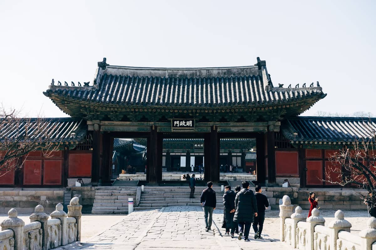 Visitantes caminham em direção ao portão de entrada do Santuário Jongmyo em Seul em um dia ensolarado, com arquitetura coreana tradicional e caminho de pedra.