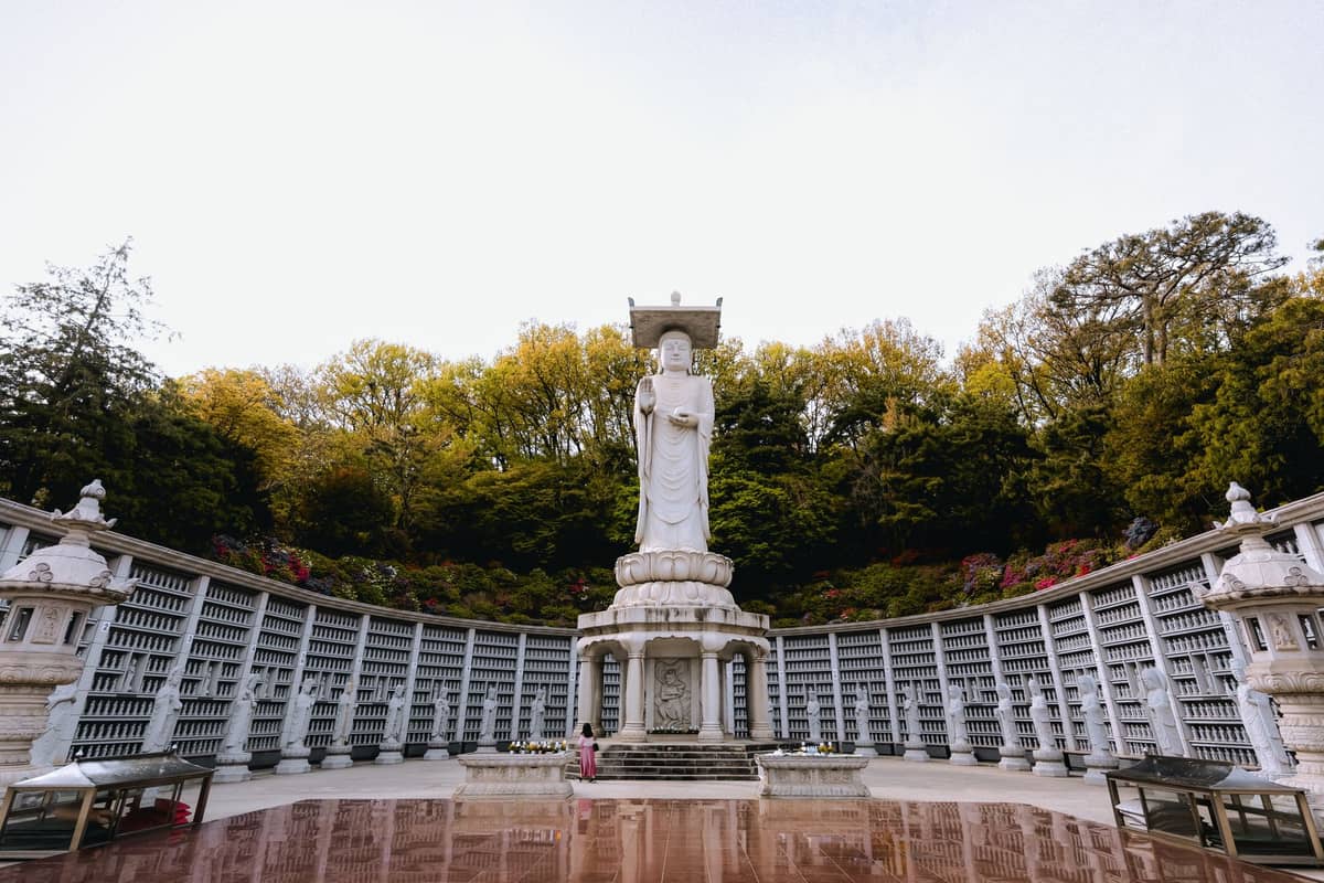 Vista ampla da enorme estátua de Buda no Templo Bongeunsa, em Seul, emoldurada por um columbário semicircular e árvores na primavera.