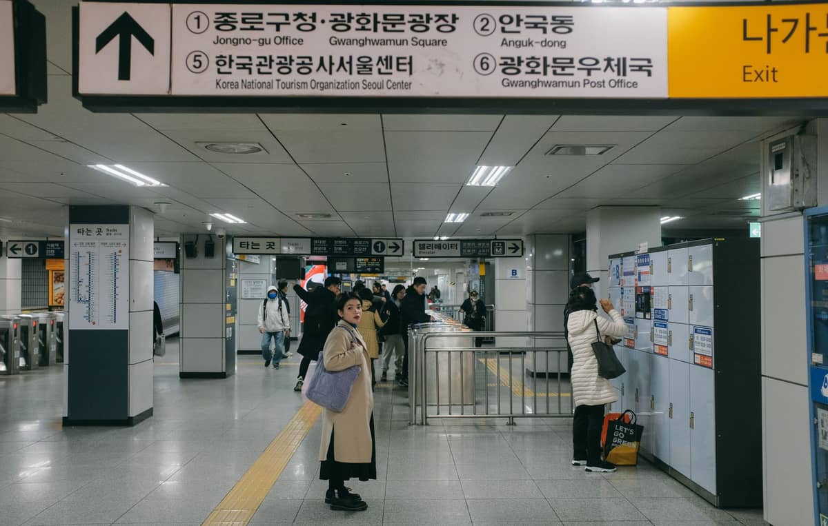 Vista interior de uma movimentada estação de metrô de Seul com passageiros, placas de direção em coreano e inglês, escadas rolantes e armários.