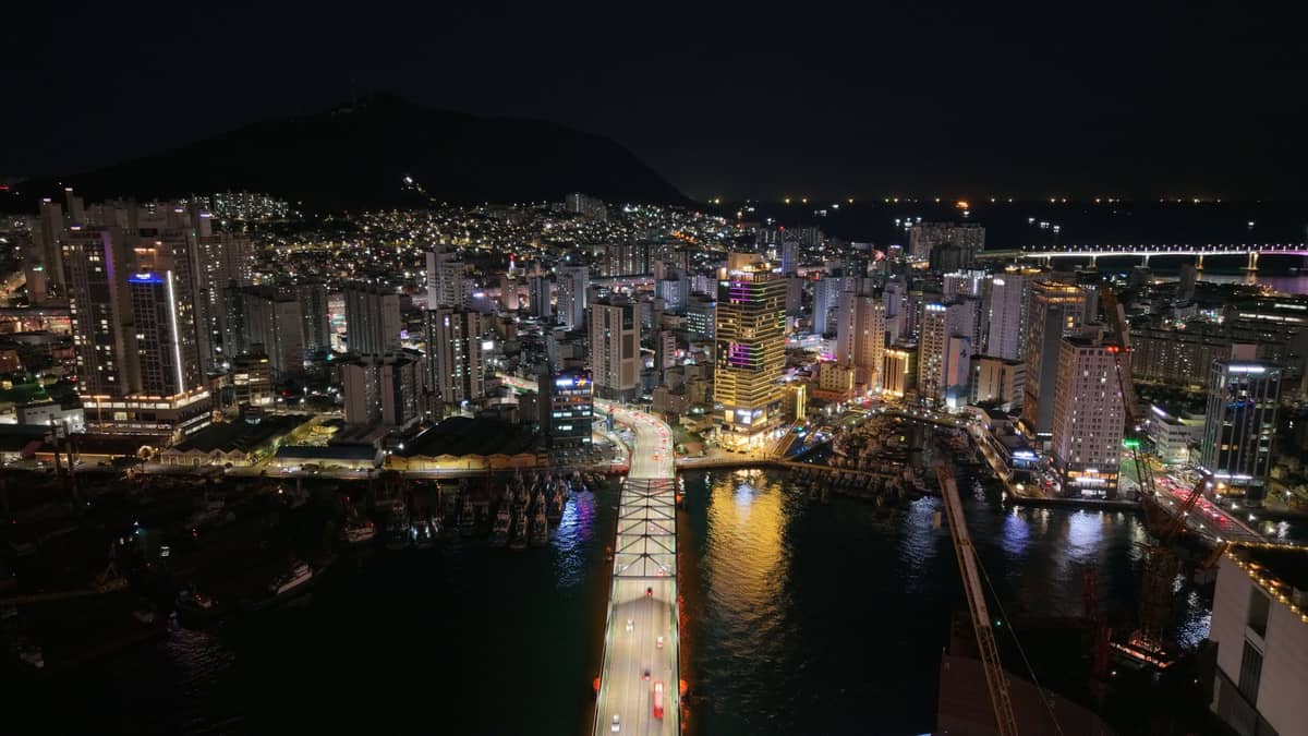 Vista aérea noturna da paisagem urbana iluminada de Busan, com uma ponte levando ao centro vibrante da cidade e morros escuros ao fundo.