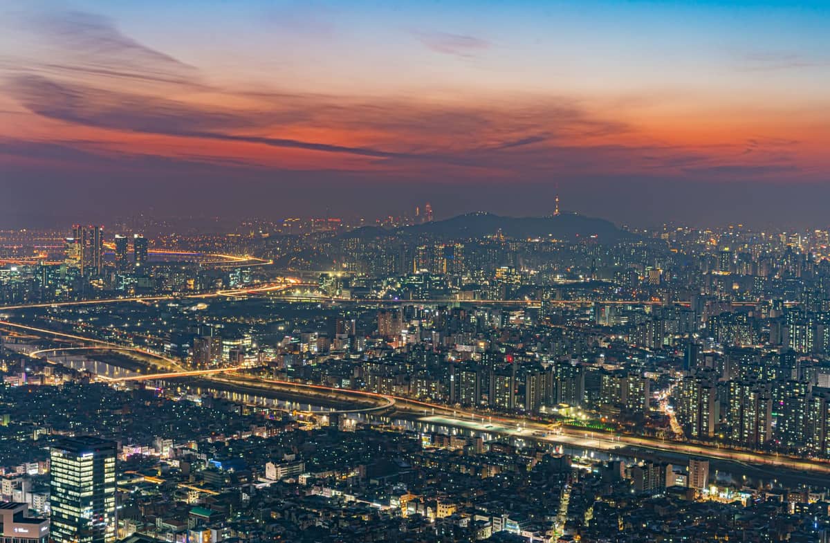 Vista panorâmica noturna de Seul com luzes da cidade, o Rio Han e a Torre Namsan Seoul sob um dramático céu ao pôr do sol.