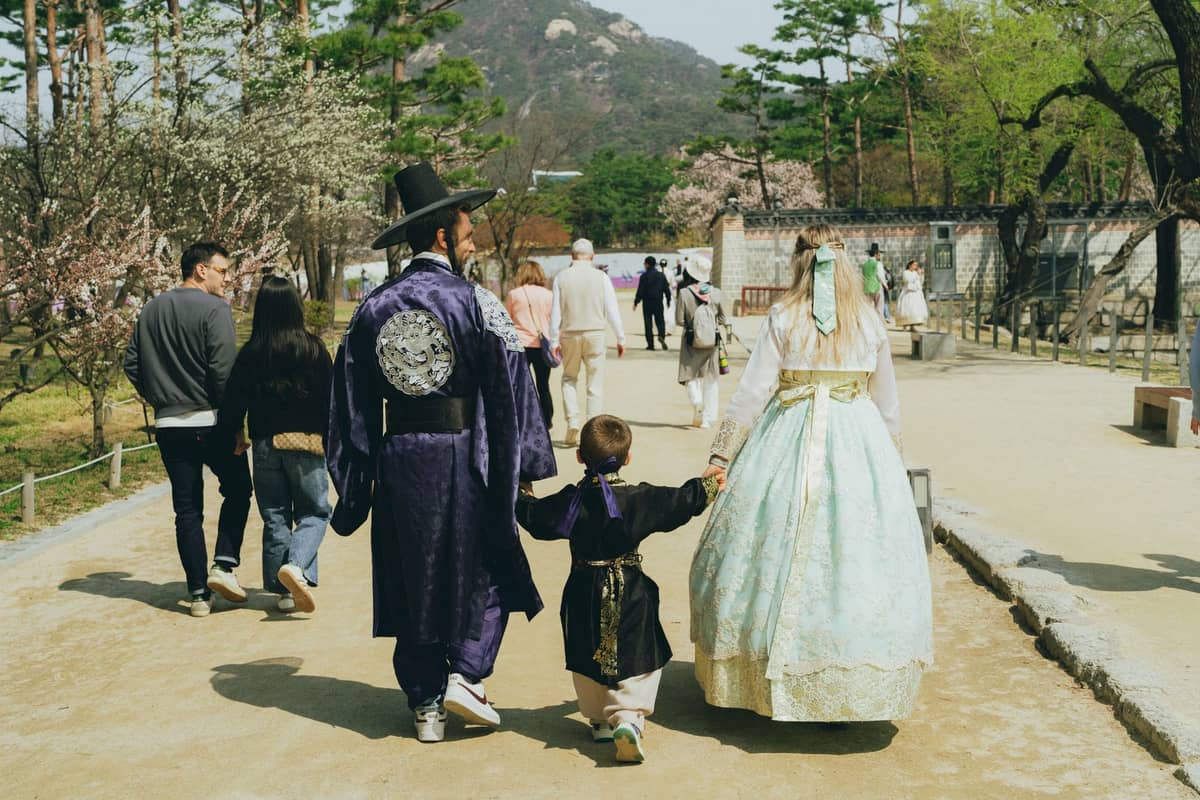 Família com uma criança vestindo hanbok tradicional caminhando juntos em um belo parque de palácio em Seul, rodeados de árvores e outros visitantes.