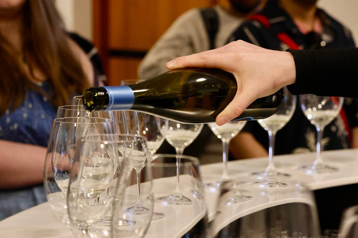 A hand pours white wine into several tasting glasses at Seppeltsfield Winery, with people in the background and warm indoor lighting.