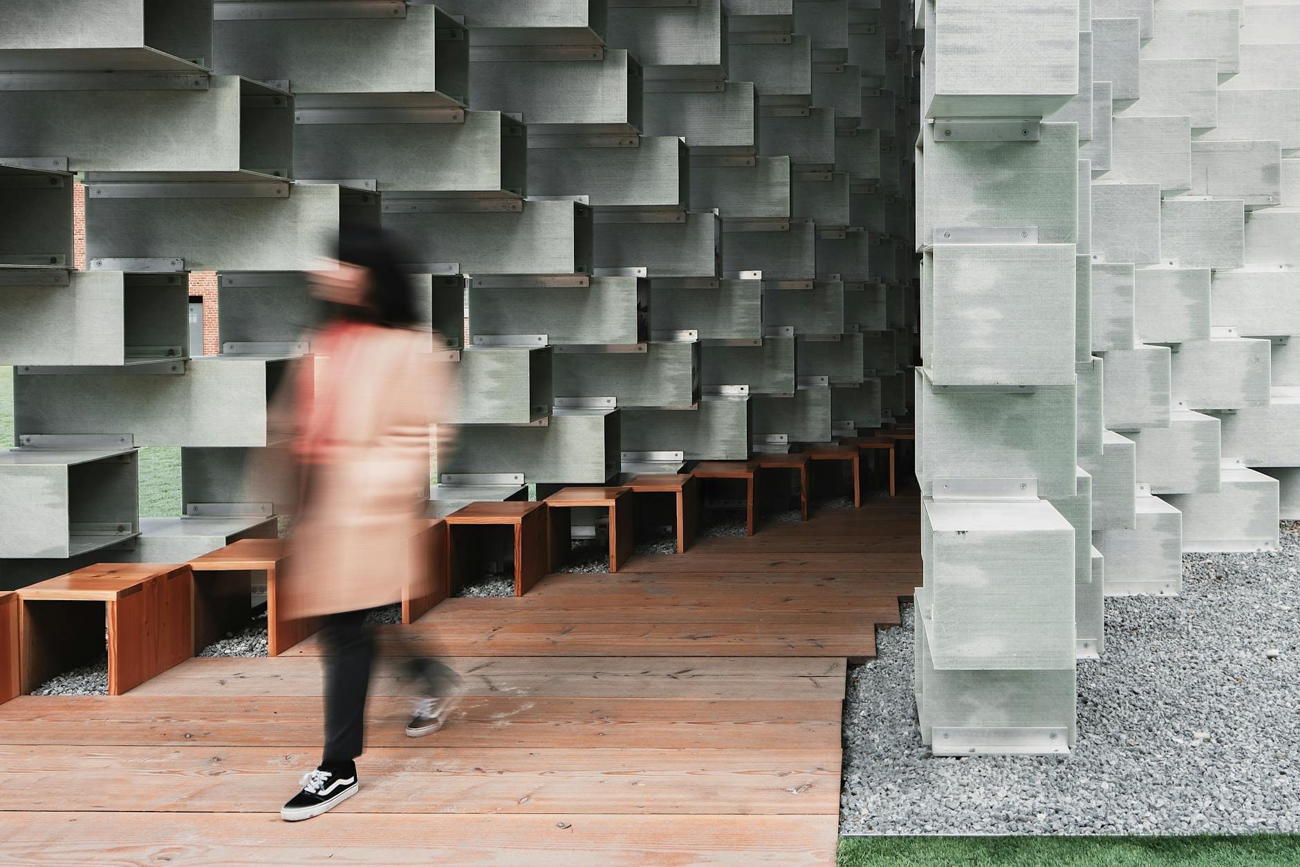 Una persona che cammina attraverso il caratteristico padiglione geometrico delle Serpentine Galleries, con dettagli architettonici moderni e luce naturale che riempie lo spazio.