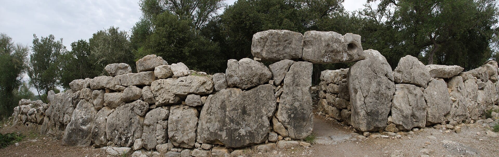 Vista panoramica delle antiche mura ciclopiche in pietra del sito archeologico di Ses Païsses circondate da lecci verdi sotto un cielo nuvoloso.