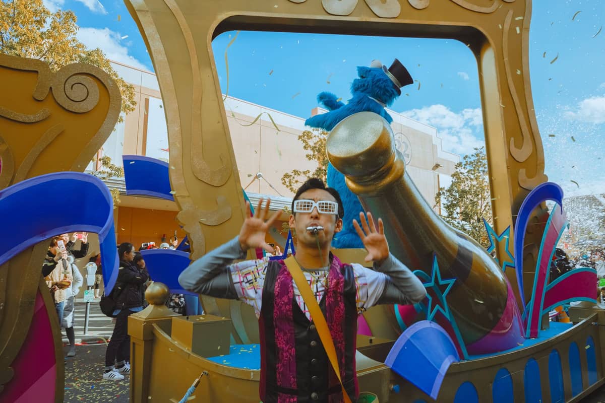 Ein Sesamstraße-Festwagen bei der Parade im Sesame Place San Diego mit einem Darsteller im Vordergrund und der Cookie-Monster-Figur im Hintergrund, umgeben von bunten Dekorationen.