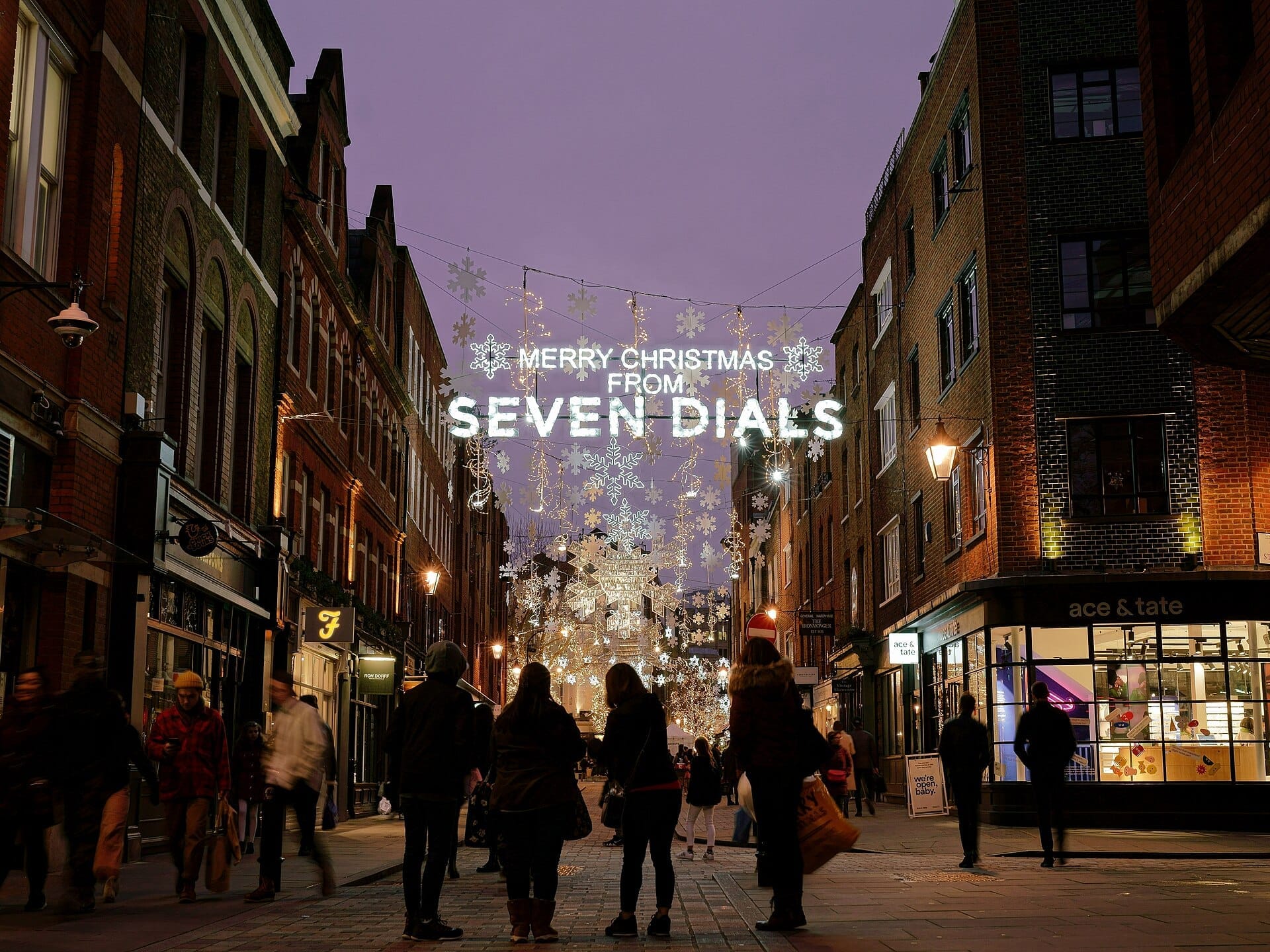Vista serale di Seven Dials a Londra con luci natalizie festive, decorazioni a fiocco di neve e persone che camminano lungo storici edifici in mattoni.