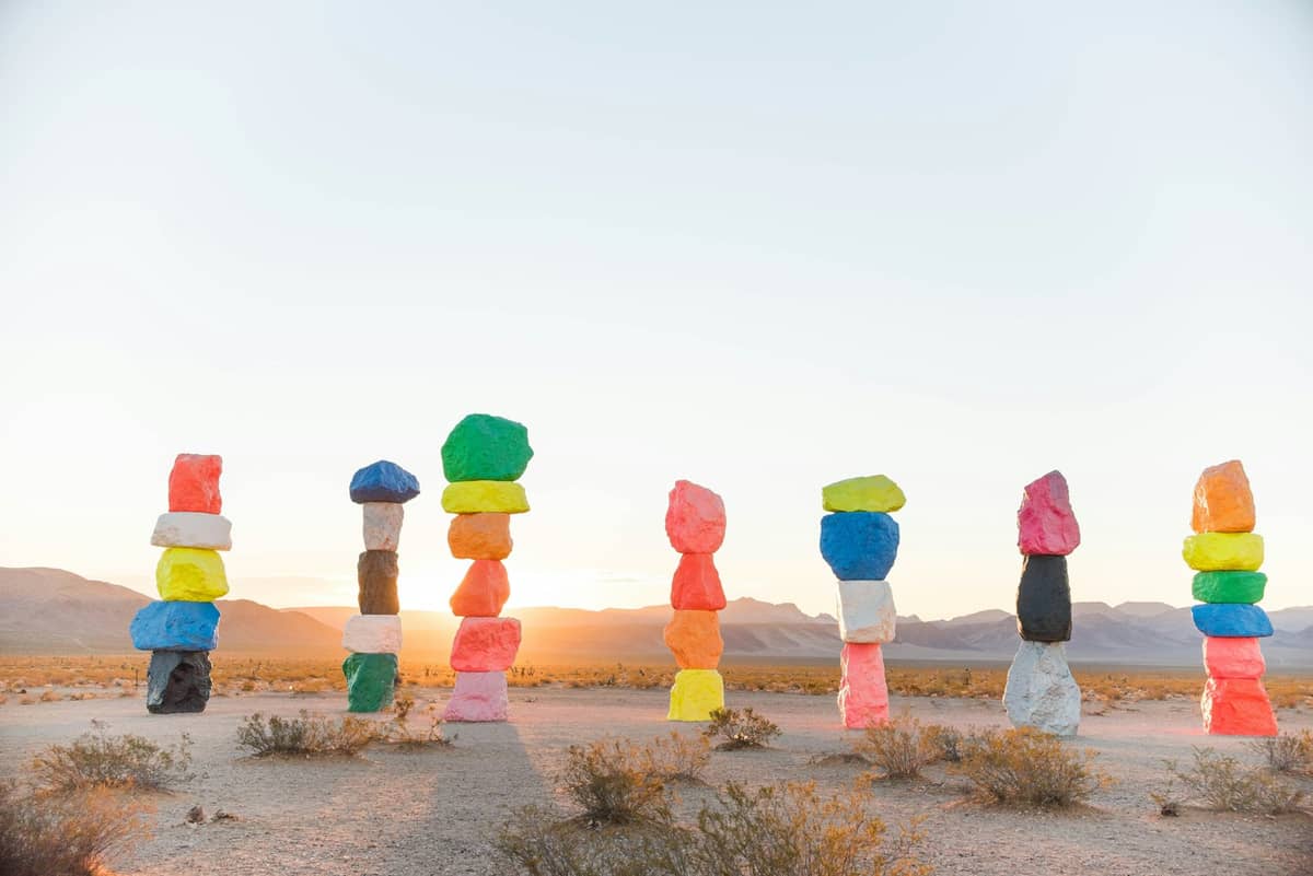 Sept grandes tours de rochers peints en couleurs néon se dressent dans le désert Mojave au lever du soleil, avec des montagnes en arrière-plan.