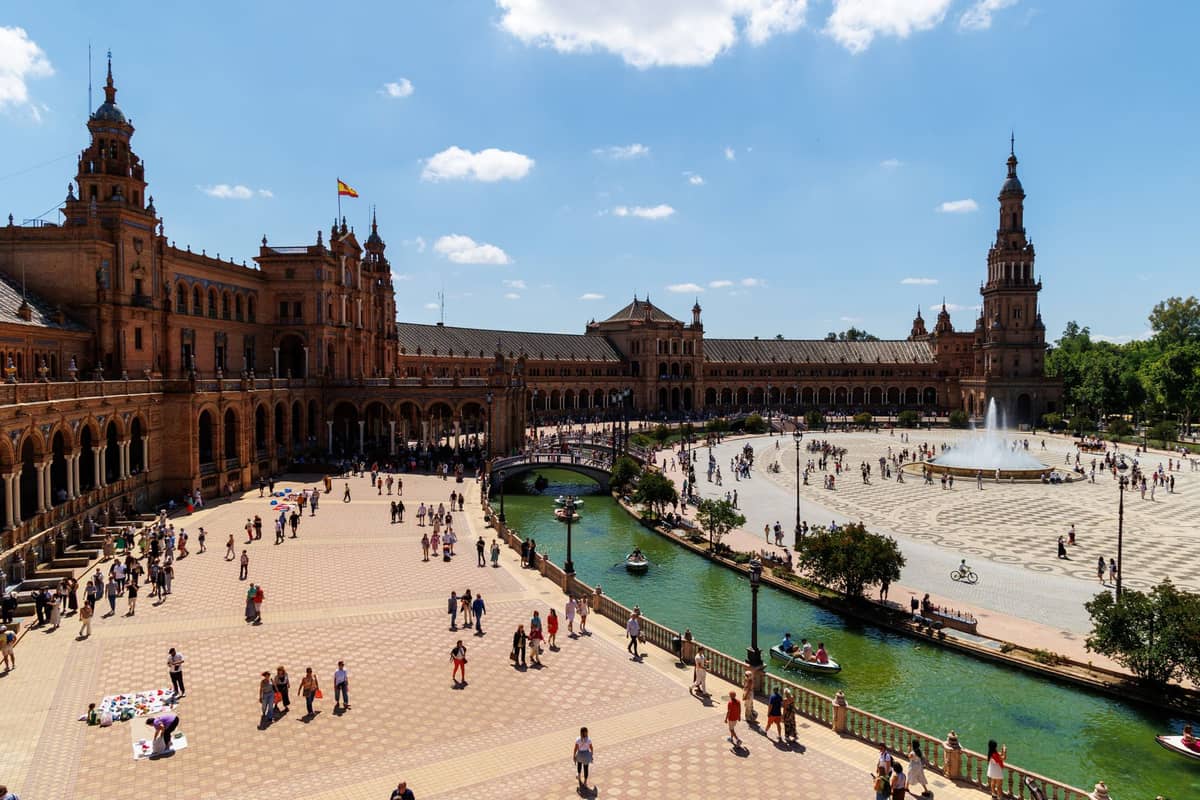วิวกว้างของ Plaza de España ในเมืองเซบิยาในวันที่แดดสวยงาม พร้อมฝูงชน คลอง และน้ำพุกลางลาน สะท้อนบรรยากาศคึกคักของปลายฤดูร้อน