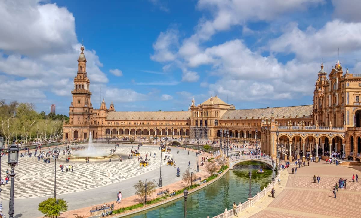 วิวกว้างของ Plaza de España ในเซบีญา สเปน ตึกโค้ง น้ำพุใหญ่ คลอง และผู้คนที่เพลิดเพลินกับแดดฤดูใบไม้ผลิ