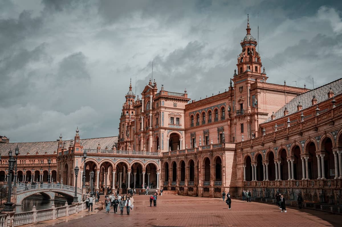 วิวกว้างของ Plaza de España ในเมือง Seville พร้อมผู้คนที่เดินบนพื้นเปียกภายใต้ท้องฟ้าฤดูใบไม้ผลิที่มืดครึ้ม