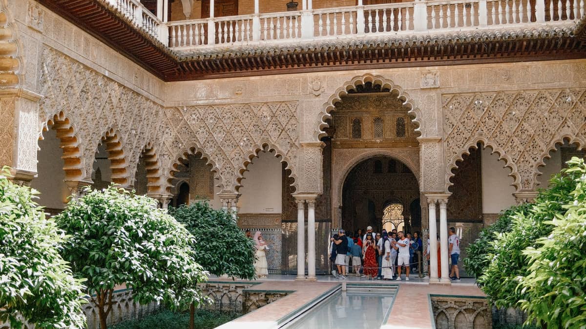นักท่องเที่ยวสำรวจลานโค้งอันเป็นสัญลักษณ์ของ Real Alcázar ในเมือง Seville ซึ่งเต็มไปด้วยต้นไม้เขียวขจีและสถาปัตยกรรมแบบมัวร์ที่งดงาม
