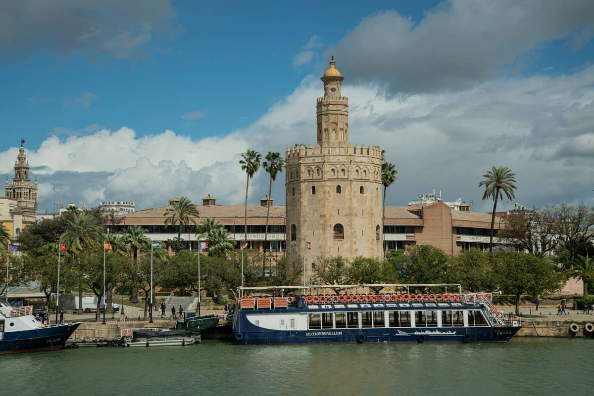 วิวทิวทัศน์ของ Torre del Oro ในเมืองเซบิยาริมฝั่งแม่น้ำภายใต้ท้องฟ้าฤดูหนาวที่มีเมฆบางส่วน พร้อมเรือที่จอดเทียบท่าและนักเดินเท้าบางส่วนอยู่ใกล้เคียง