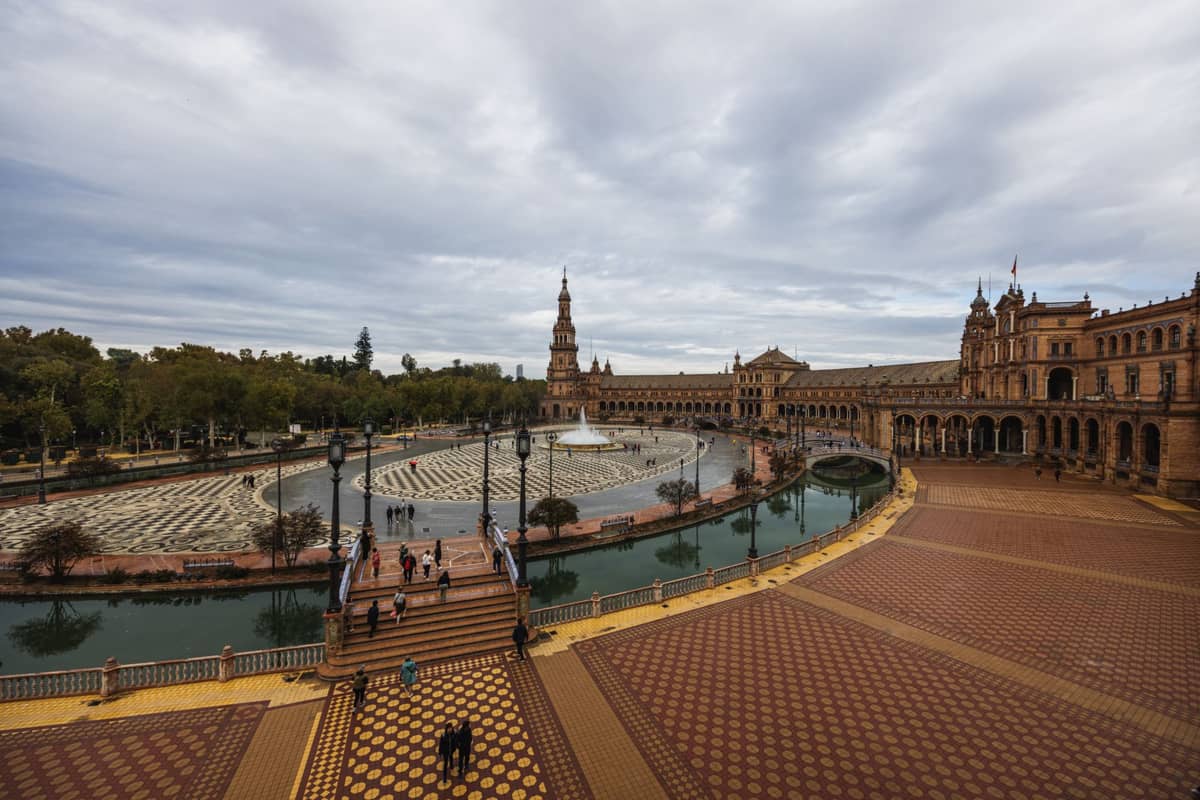 วิวกว้างของ Plaza de España ในเซบียาช่วงฤดูหนาววันที่ฟ้าครึ้ม แสดงลานโค้ง หอคอย และคลอง มีนักท่องเที่ยวน้อย