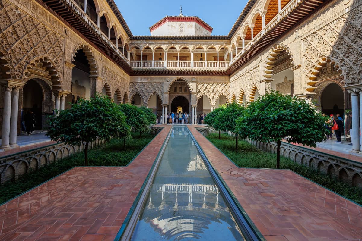 วิวทิวทัศน์ของลานกลางอันโด่งดังของ Real Alcázar ในเมือง Seville พร้อมกำแพงโค้งโค้ง การตกแต่งที่ประณีต และสระน้ำสะท้อนแสงตรงกลาง เหมาะสำหรับบทความท่องเที่ยวแบบประหยัด