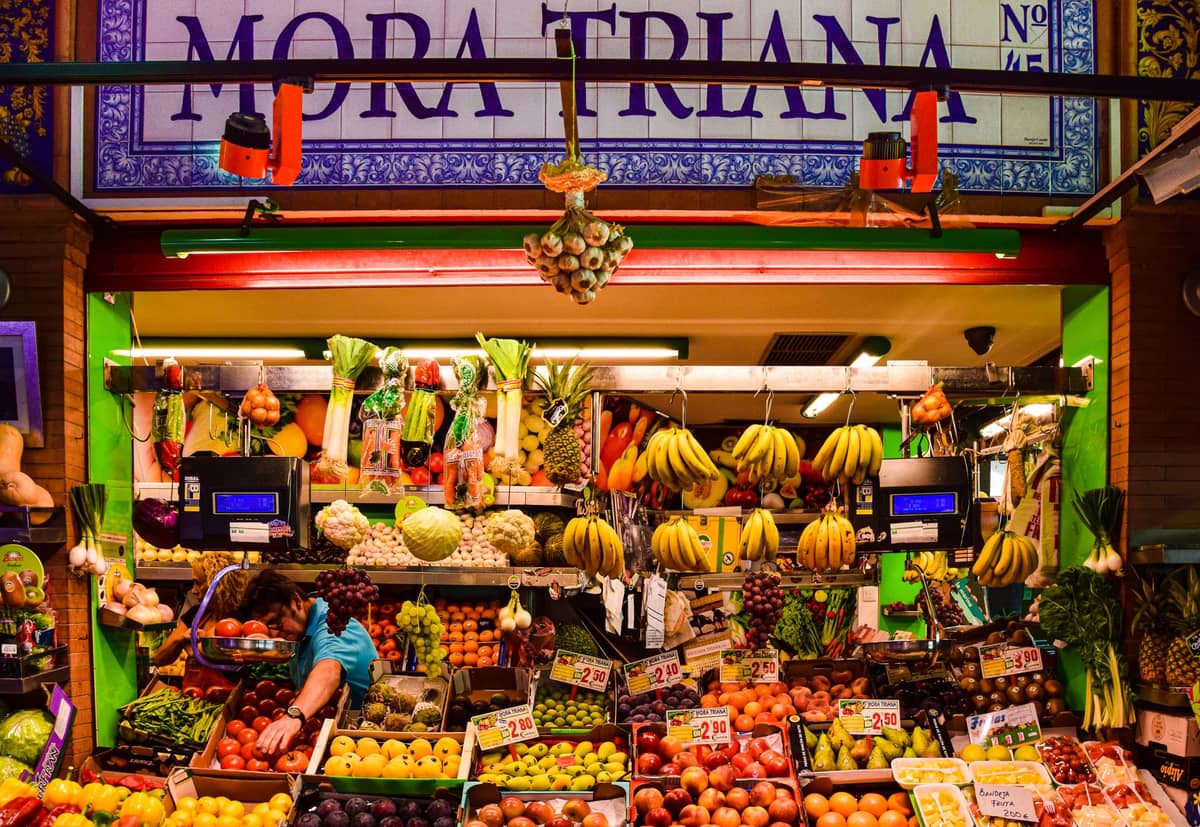 แผงตลาดสีสันสดใสพร้อมผลไม้และผักสดภายใต้ป้าย 'Mora Triana' ที่ตลาด Triana เมือง Seville
