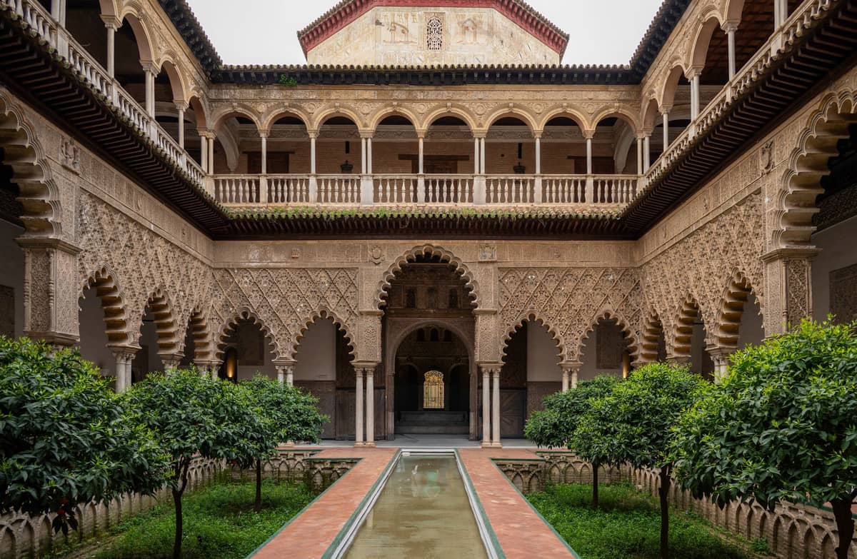ลานสวยงามของ Real Alcázar เมืองเซบียา พร้อมซุ้มโค้งสไตล์มัวร์ ต้นส้ม และน้ำพุกลางลานใต้ท้องฟ้าครึ้ม