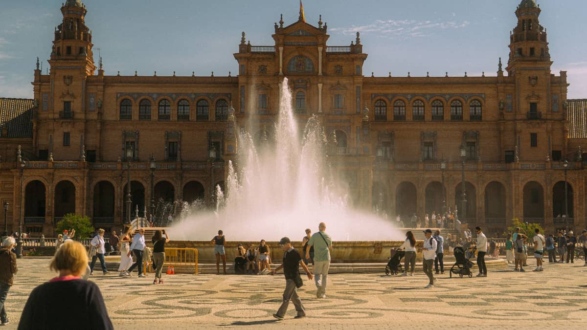 ผู้คนและครอบครัวหลายครอบครัวพร้อมรถเข็นเด็กเดินเล่นรอบๆ น้ำพุที่ Plaza de España ในเมืองเซบิยาในวันที่แดดออก