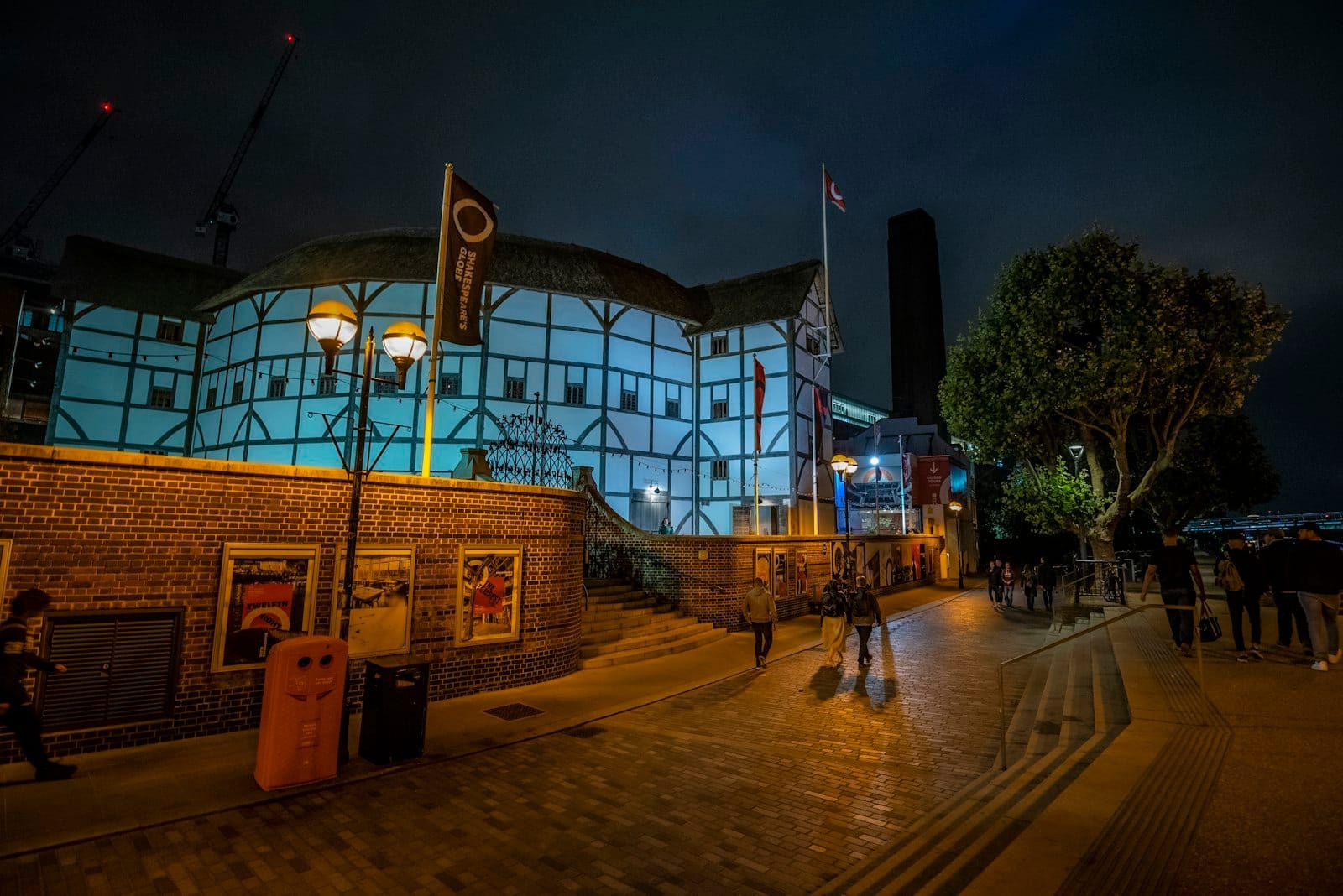 Vista esterna dello Shakespeare's Globe Theatre di Londra di notte, illuminato con luci blu, con persone che camminano lungo il percorso illuminato davanti al teatro.