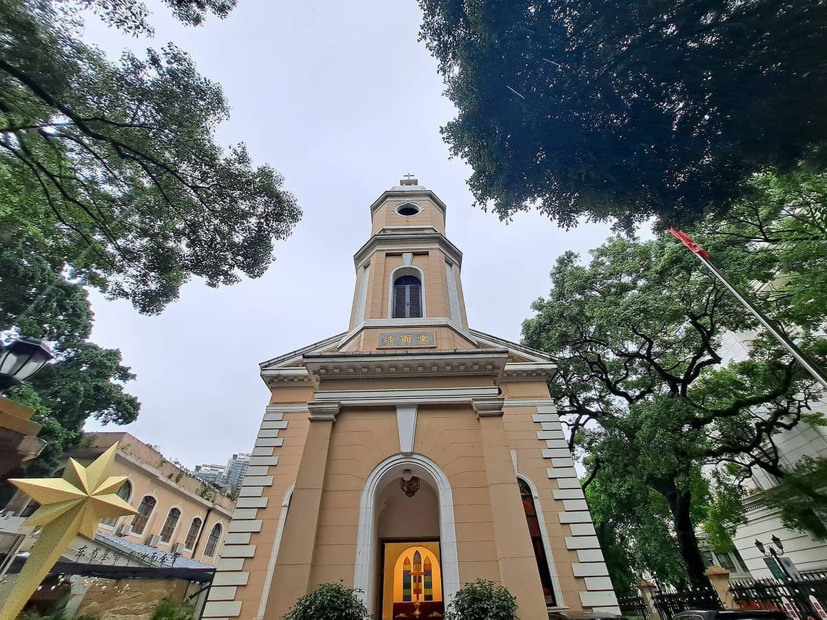 Front view of Shamian Christian Church with its tan façade, arched entrance, and bell tower framed by leafy green trees.