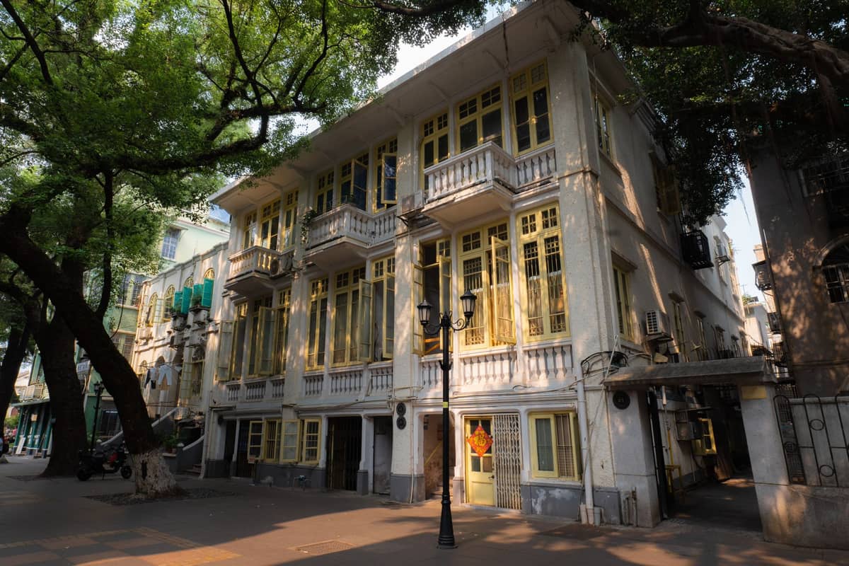 European-style historic building with yellow windows and balconies, shaded by large trees on Shamian Island's peaceful, leafy street in Guangzhou.