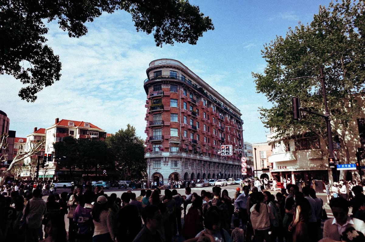 Historischer Blick auf Shanghais ikonisches Wukang Mansion an einer belebten Kreuzung, mit Menschenmassen und belaubten Bäumen, als Beispiel kolonialer Architektur.