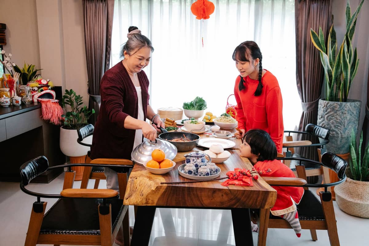 Tre membri della famiglia riuniti attorno a un tavolo da pranzo in legno coperto di piatti tradizionali cinesi e decorazioni per il Festival di Primavera.
