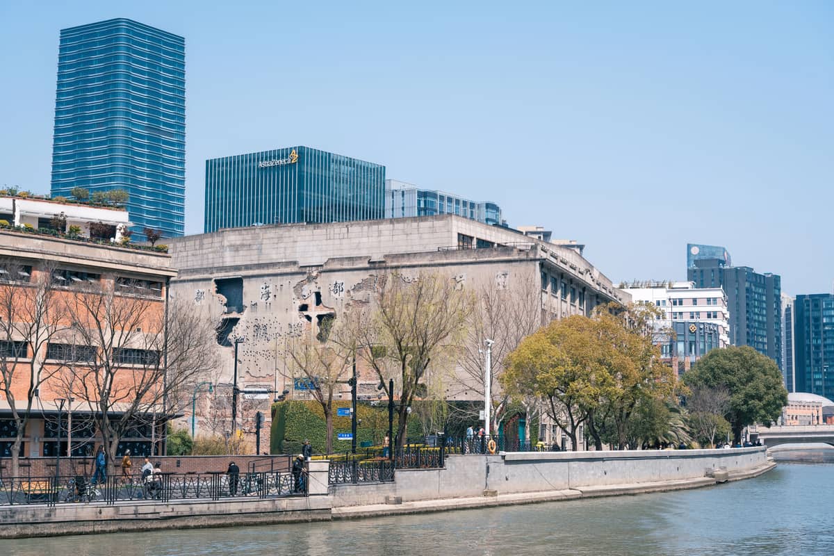 Rue pittoresque en bord de canal à Shanghai avec des piétons, des arbres, un bâtiment industriel historique et des gratte-ciel en verre modernes en arrière-plan.