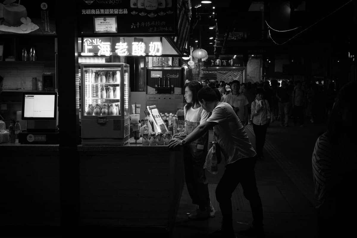 Black and white scene of a night market in Shanghai with people at a drink stall and illuminated signage.