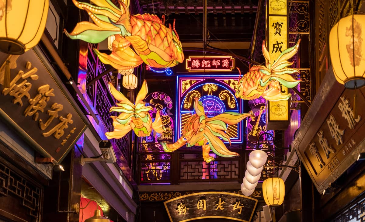 Colorful lanterns and neon signs illuminate a narrow traditional street inside Yu Garden commercial zone, capturing Old Town Shanghai night market atmosphere.