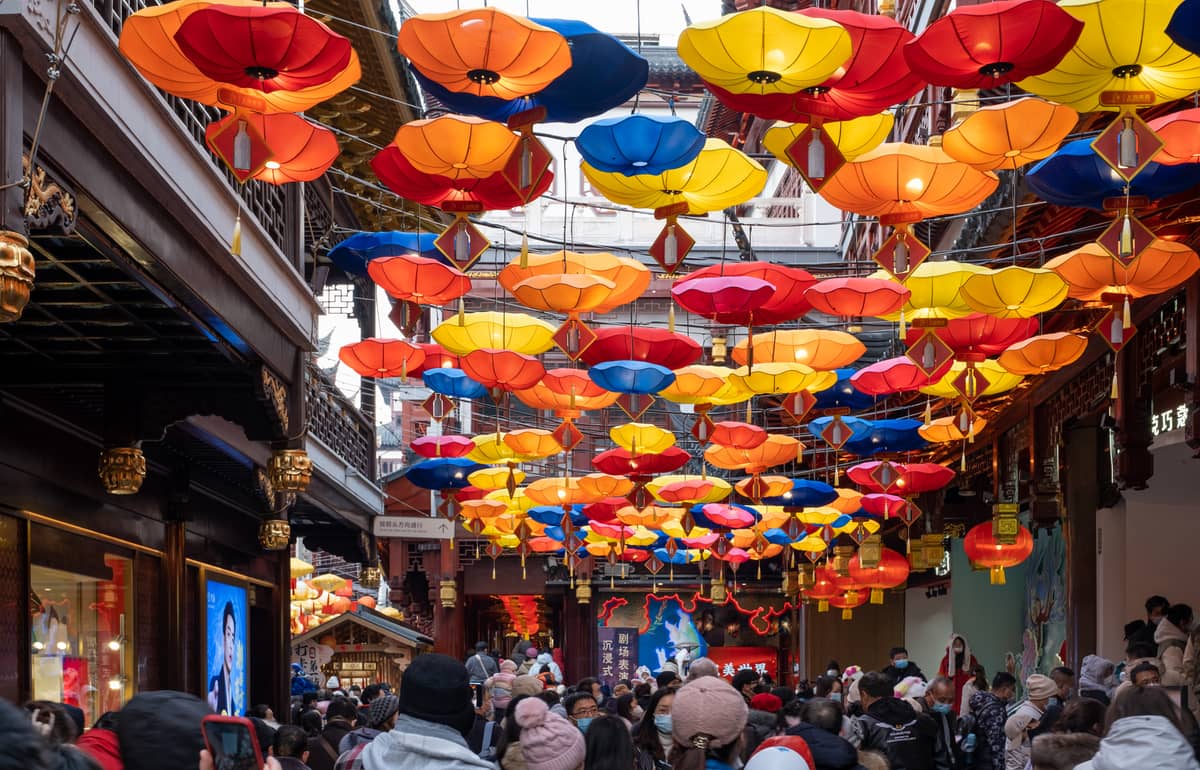 Belebte Essensstraße in Shanghai, geschmückt mit bunten Laternen, umgeben von traditioneller Architektur und voller Menschen.