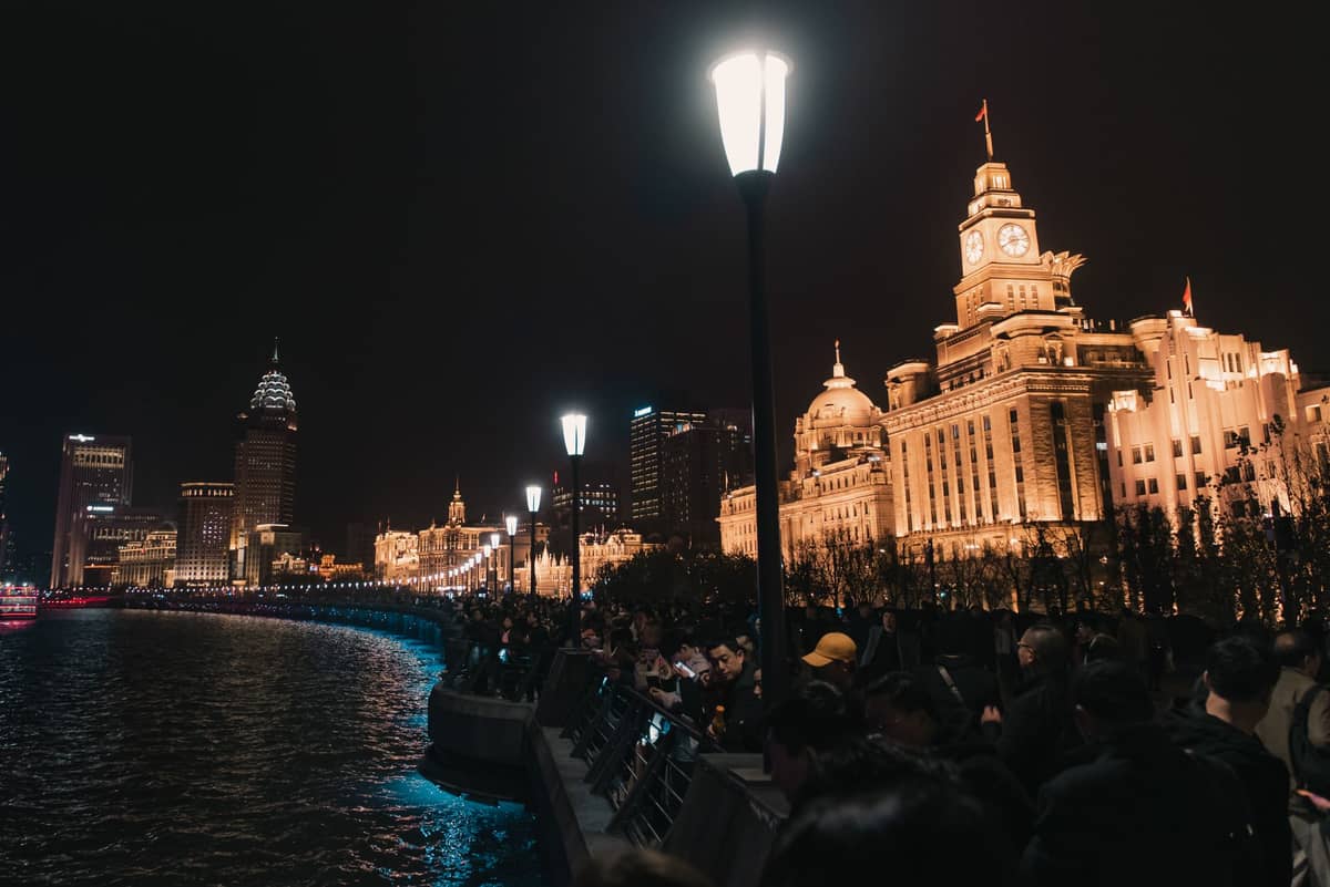 Night view of the Bund in Shanghai with crowds of people walking along the riverfront promenade, street lamps, and historic buildings brightly lit.