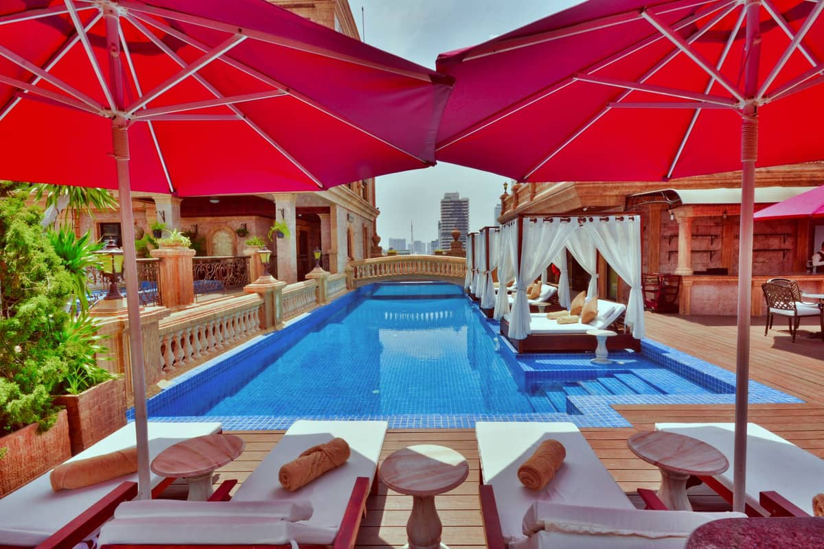 Luxuriöser Dachpool mit roten Sonnenschirmen, Liegestühlen und der Stadtsilhouette in der Ferne, der Entspannung in einem gehobenen Hotel in urbaner Umgebung andeutet.