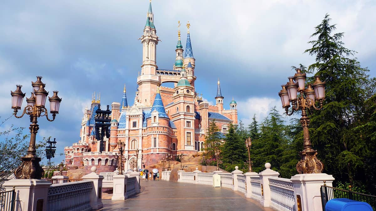 Weitwinkelansicht des Enchanted Storybook Castle im Shanghai Disneyland, mit verzierten Laternen, einer Brücke und üppigem Grün unter blauem Himmel.