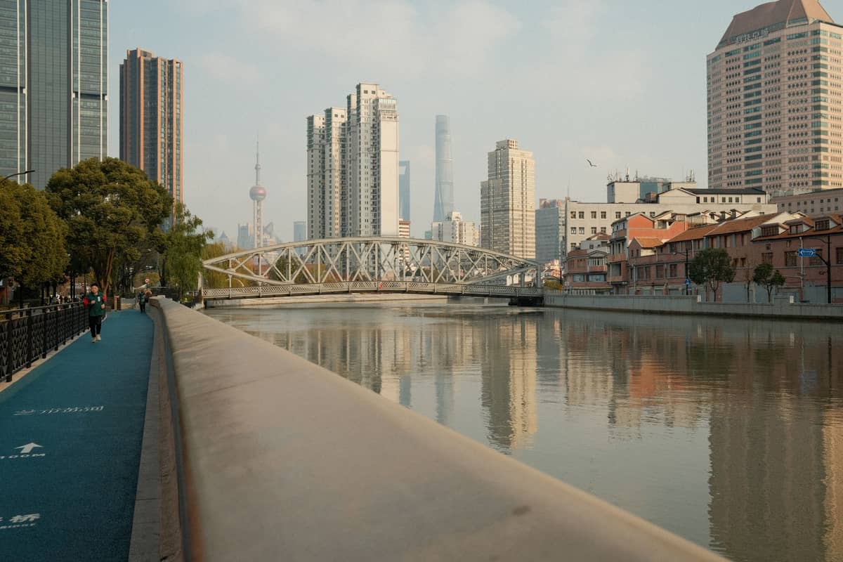Malerischer Tagesblick auf die Skyline von Shanghai mit Bäumen, einem blauen Flussuferweg, modernen Gebäuden und dem Oriental Pearl Tower in der Ferne.